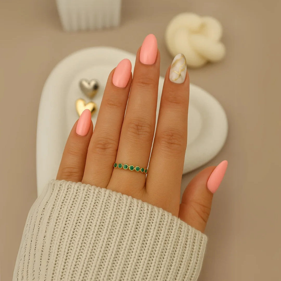 Mano femenina con uñas largas color rosa y diseño mármol, usando un anillo dorado con piedras verdes, sobre superficie de tela blanca y fondo decorativo en tonos neutros.
