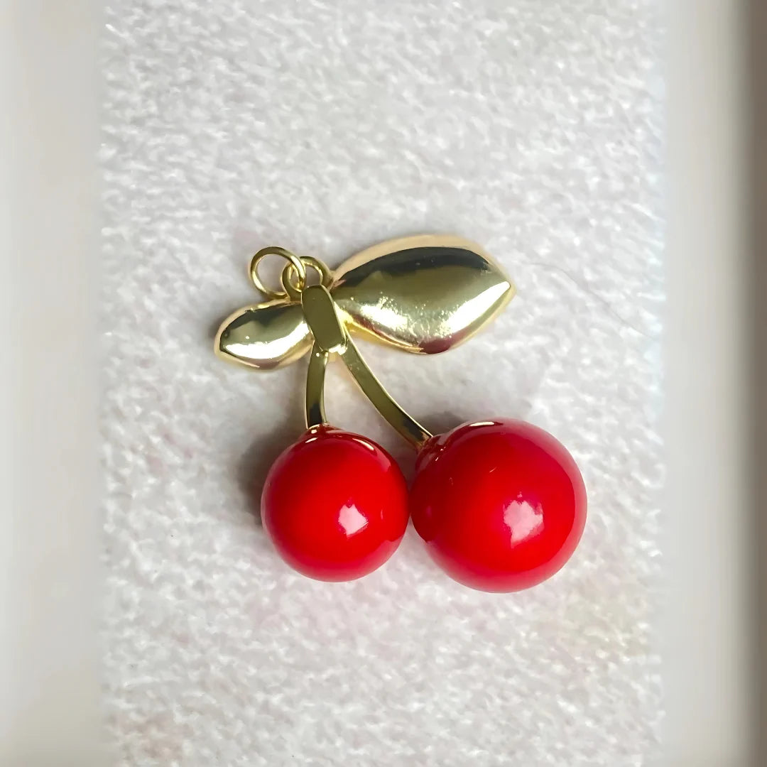 Colgante de cerezas rojas con acabado dorado sobre superficie blanca

