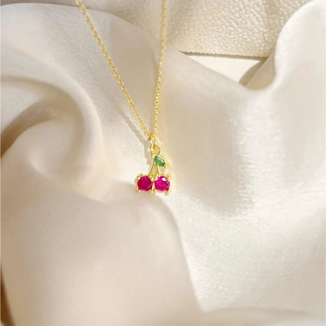 Collar cherry con cereza esmaltada rosada y cadena dorada sobre fondo beige

