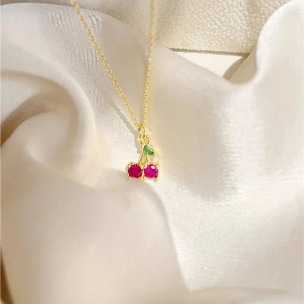 Collar cherry con cereza esmaltada rosada y cadena dorada sobre fondo beige

